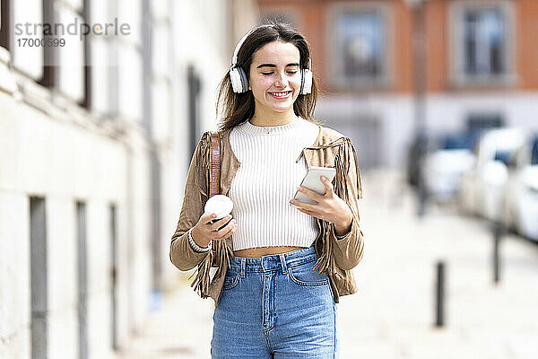 Lächelnde Frau mit Kopfhörern und Smartphone auf dem Fußweg