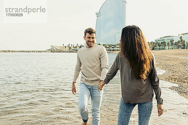 Lächelndes Paar hält sich an den Händen  während es am Strand über das Wasser läuft