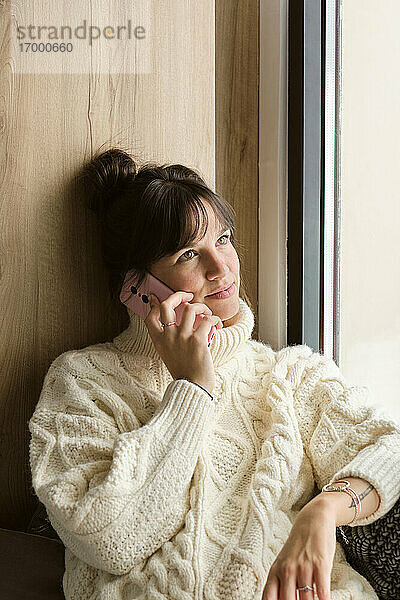 Schöne Frau im Pullover  die aufschaut  während sie am Fenster zu Hause mit dem Handy telefoniert