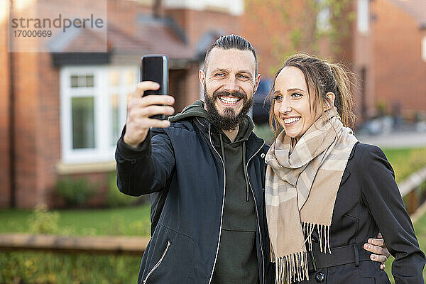 Lächelnder Mann  der ein Selfie mit seiner Freundin macht  während er vor seinem neuen Haus steht