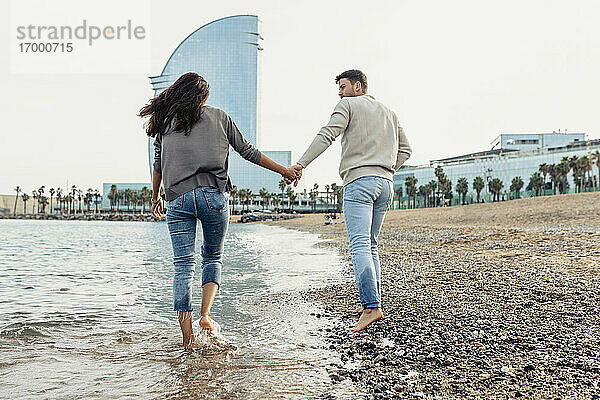 Freundin und Freund halten sich an den Händen  während sie am Strand im Wasser spazieren gehen