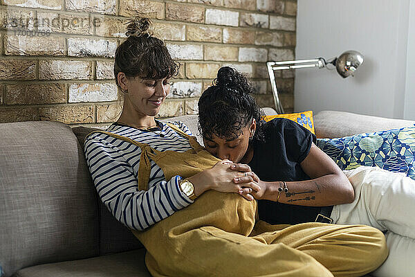 Frau küsst schwangere Freundin Bauch beim Sitzen auf Sofa zu Hause