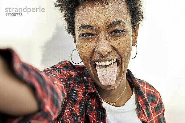 Fröhliche junge Frau  die ein Selfie macht  während sie ihre Zunge gegen eine weiße Wand streckt