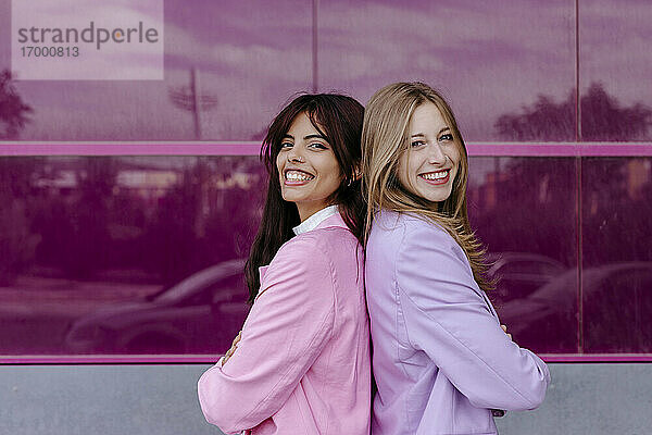 Lächelnde Schwestern stehen Rücken an Rücken vor einer Acrylglaswand