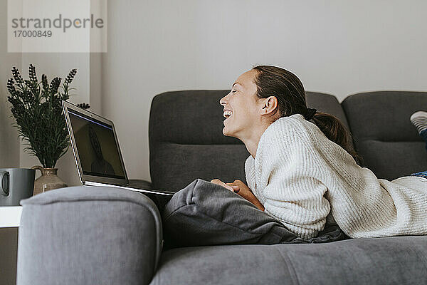 Frau lacht  während sie mit einem Freund über einen Videoanruf am Laptop zu Hause spricht