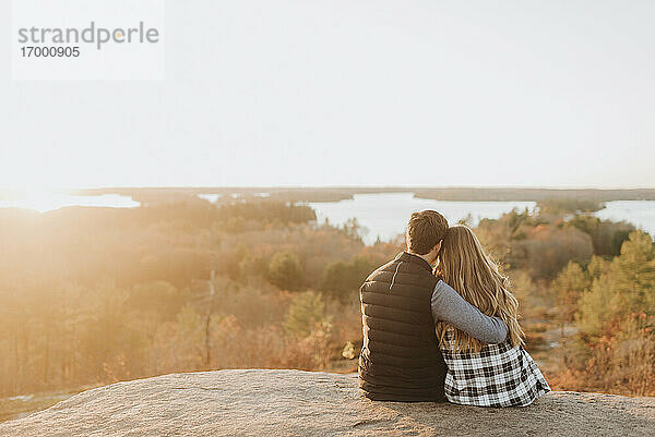 Junges Paar sitzt zusammen auf einem Felsen und bewundert die untergehende Sonne im Herbst