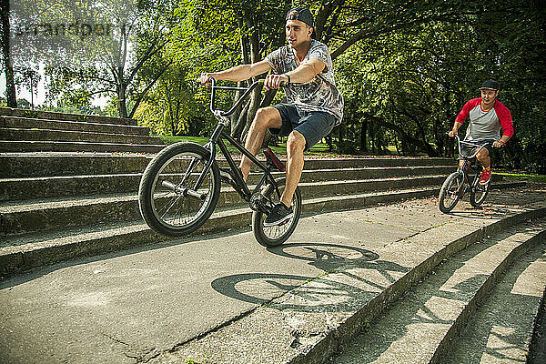 Junger Mann macht Wheelie-Stunt mit BMX-Fahrrad gegen einen Freund  der auf den Stufen eines öffentlichen Parks radelt