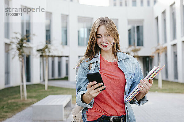 Lächelnde junge blonde Studentin  die ein Smartphone an der Universität benutzt