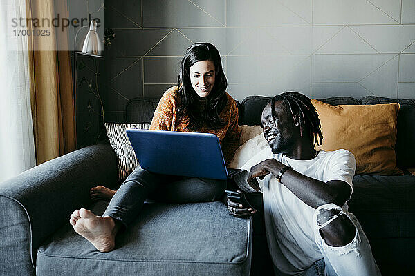 Lächelnder junger Mann mit schwangerer Frau  die einen Laptop benutzt  während sie zu Hause auf dem Sofa sitzen
