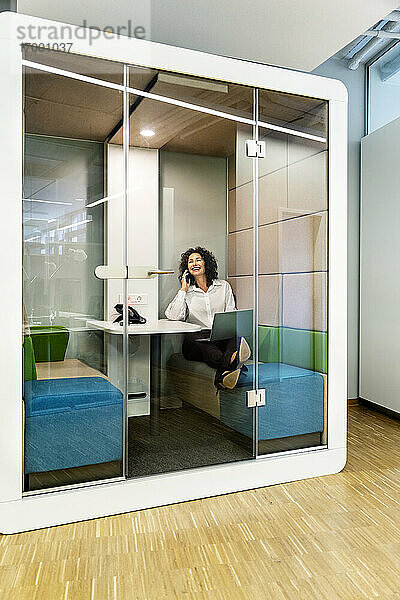 Geschäftsfrau mit Laptop  die in einer Kabine im Büro sitzt und mit ihrem Mobiltelefon spricht
