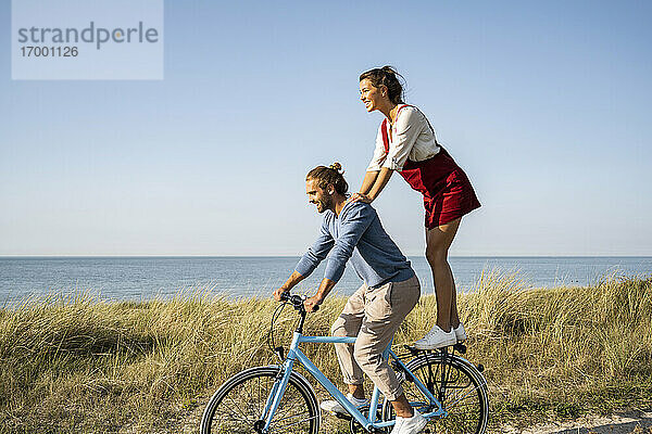 Lächelnde Frau  die eine Fahrt mit einem Mann genießt  während sie auf einem Fahrrad gegen den klaren Himmel steht
