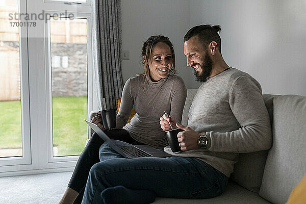 Lächelndes junges Paar trinkt Tee und benutzt einen Laptop auf dem Sofa zu Hause