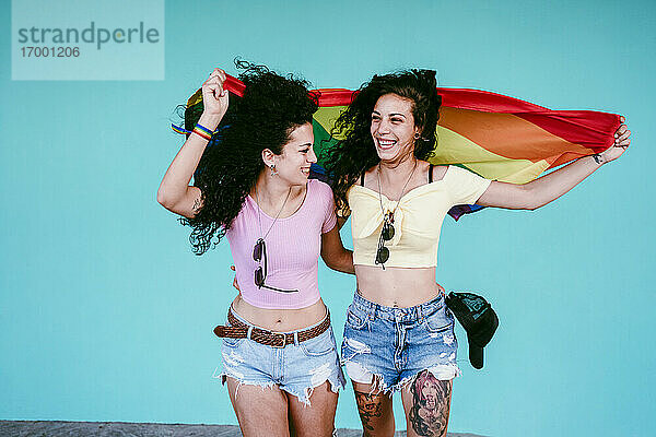 Fröhliche lesbische Freundinnen halten eine Regenbogenfahne  während sie an der Wand stehen