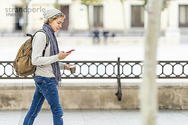 Junger Mann mit Einwegbecher und Mobiltelefon beim Spaziergang in der Stadt