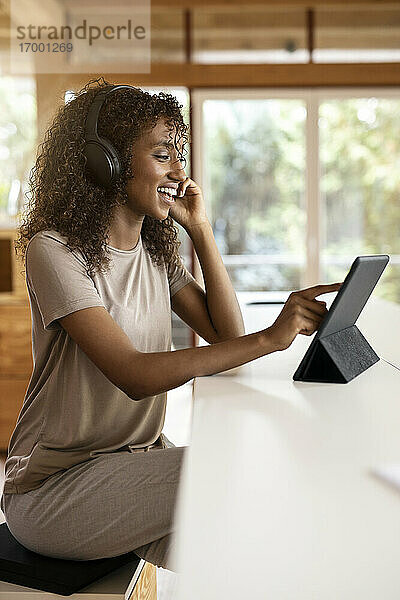 Lächelnde Geschäftsfrau mit digitalem Tablet  die über Kopfhörer spricht  während sie zu Hause sitzt