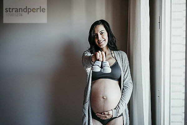 Lächelnde schwangere Frau zeigt Baby-Booties  während sie zu Hause an der Wand steht