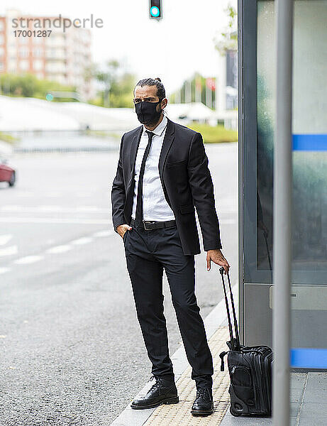 Geschäftsmann mit Gesichtsschutzmaske  der mit einem Koffer an der Bushaltestelle wartet