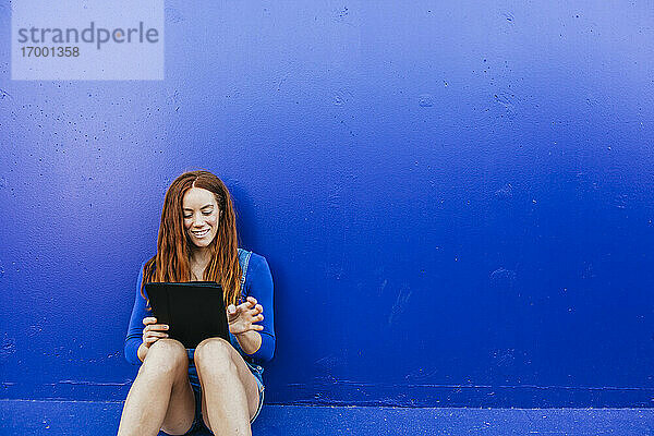 Junge Frau benutzt ein digitales Tablet  während sie an einer blauen Wand sitzt