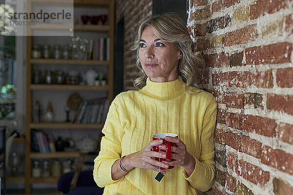 Reife Frau mit Teetasse  die wegschaut  während sie sich an eine Backsteinmauer in der Küche lehnt