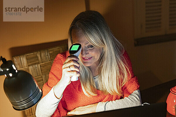Lächelnde ältere Frau  die ein Infrarot-Thermometer benutzt  während sie einen Videoanruf auf einem Laptop zu Hause betrachtet