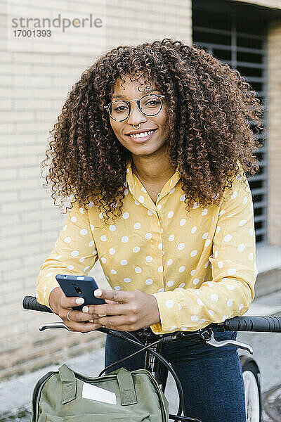 Lächelnde Frau  die ein Mobiltelefon benutzt  während sie mit dem Fahrrad an der Wand steht