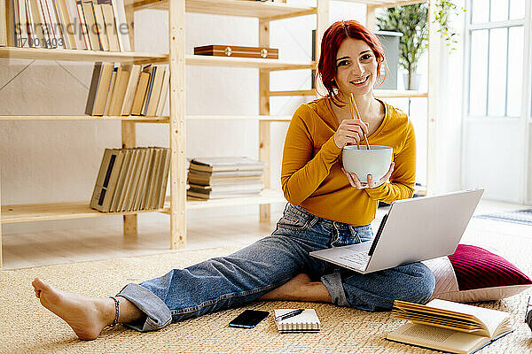 Lächelnde Frau beim Essen und Lernen am Laptop zu Hause sitzend