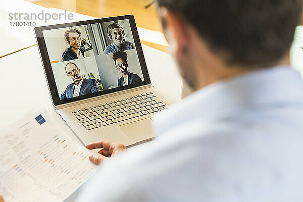 Geschäftsmann hält Papier  während er an einer Videokonferenz mit einem Team auf einem Laptop im Büro teilnimmt