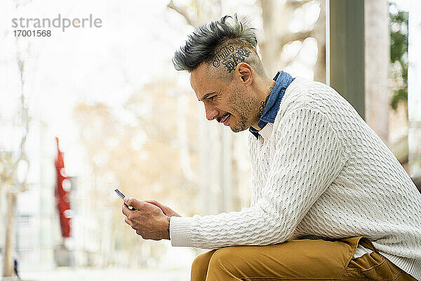 Lächelnder Geschäftsmann  der ein Mobiltelefon benutzt  während er an der Bushaltestelle sitzt