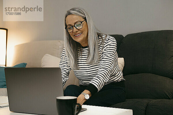 Lächelnde ältere Frau  die einen Laptop benutzt  während sie zu Hause auf dem Sofa sitzt