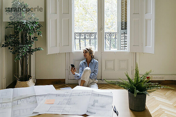 Geschäftsfrau  die ein Smartphone benutzt  während sie auf dem Boden vor dem Fenster im Büro sitzt