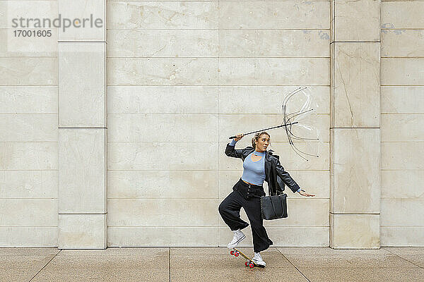 Junge Geschäftsfrau hält einen beschädigten Regenschirm  während sie an einem regnerischen Tag mit dem Skateboard gegen die Wand fährt