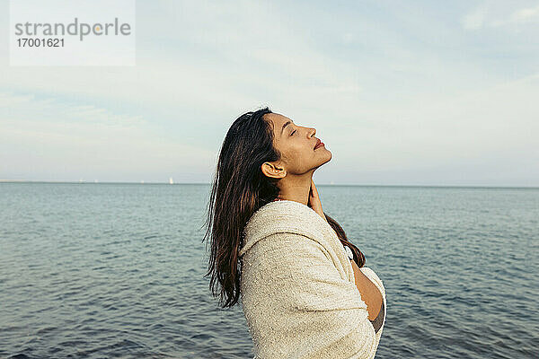 Junge Frau mit geschlossenen Augen steht gegen den Himmel am Strand