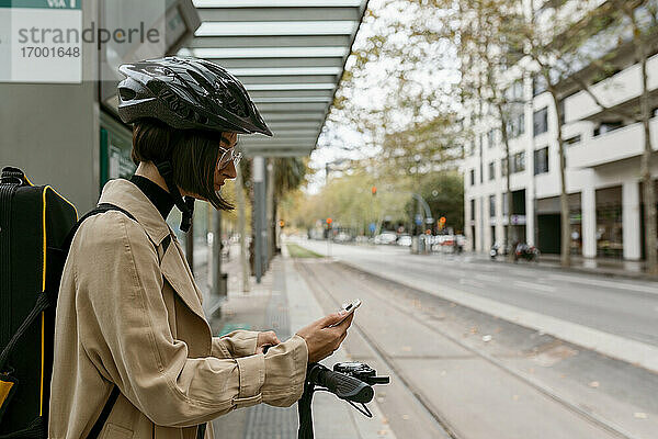 Mittlere erwachsene Frau mit Instrumentenkoffer und Elektroroller  die ein Mobiltelefon benutzt  während sie an einer Straßenbahnhaltestelle in der Stadt steht