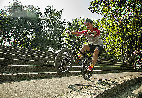 Junger asiatischer Mann macht Wheelie-Stunt mit BMX-Fahrrad auf Stufen im öffentlichen Park