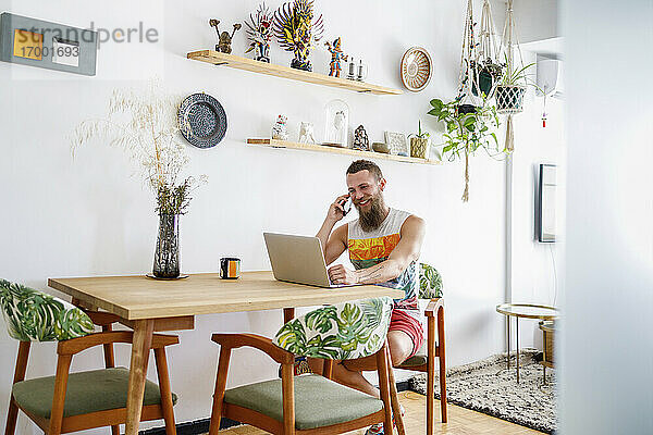 Mittlerer erwachsener Mann  der mit seinem Smartphone spricht  während er im Wohnzimmer am Laptop sitzt