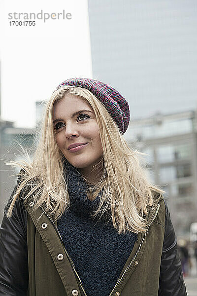 Lächelnde blonde Frau mit Strickmütze schaut weg  während sie in der Stadt steht