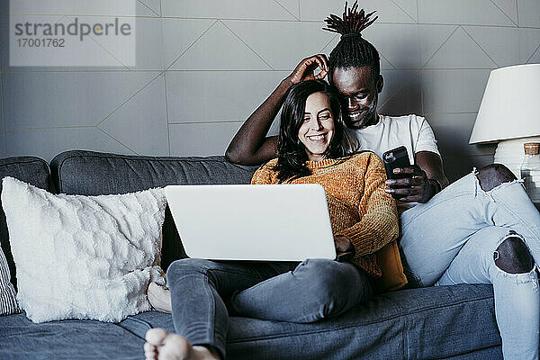Glücklicher junger Mann mit schwangerer Frau  die auf ihr Mobiltelefon schaut  während sie zu Hause einen Laptop benutzen