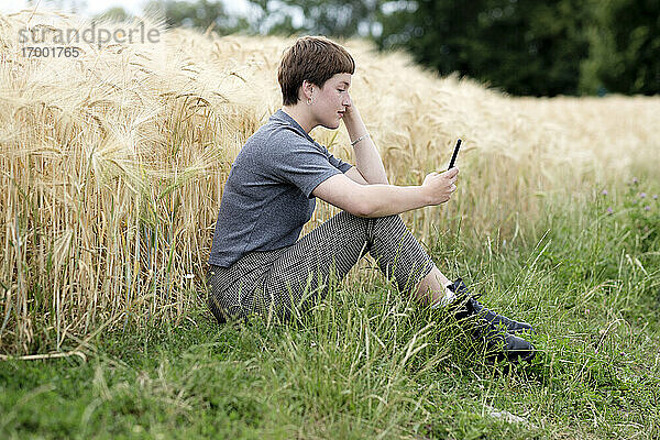 Junge Frau benutzt ihr Smartphone  während sie auf einem Gerstenfeld sitzt