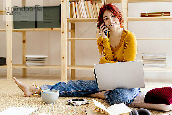 Lächelnde Frau  die mit dem Handy telefoniert  während sie zu Hause am Laptop sitzt