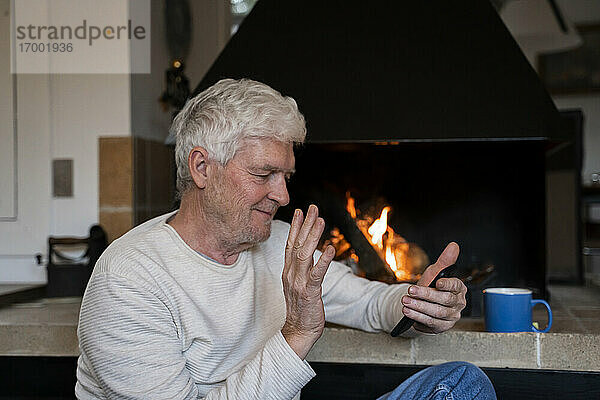 Älterer Mann winkt mit der Hand zu einem Videoanruf auf einem Mobiltelefon  während er zu Hause sitzt
