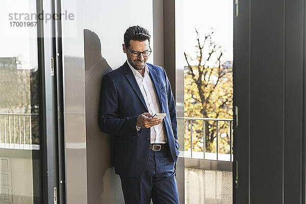 Männlicher Unternehmer  der ein Mobiltelefon benutzt  während er mit der Hand in der Tasche auf dem Balkon eines Büros steht