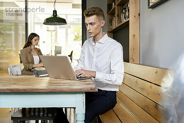 Geschäftsmann arbeitet am Laptop  während er mit einem Kollegen im Hintergrund im Büro sitzt