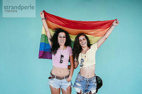 Fröhliches lesbisches Paar  das eine Regenbogenfahne hält und an der Wand steht