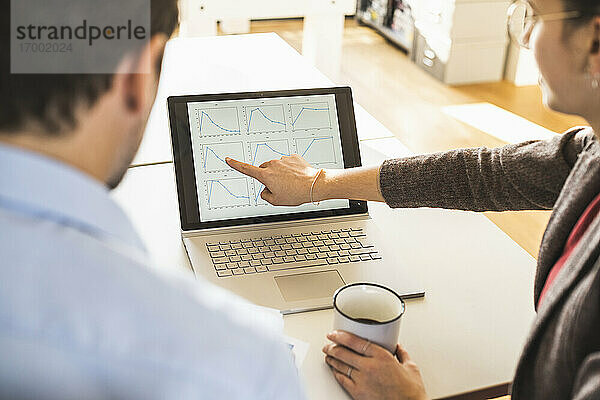 Geschäftsfrau mit Kaffeetasse  die einem Geschäftsmann im Büro sitzend ein Diagramm auf dem Laptop erklärt