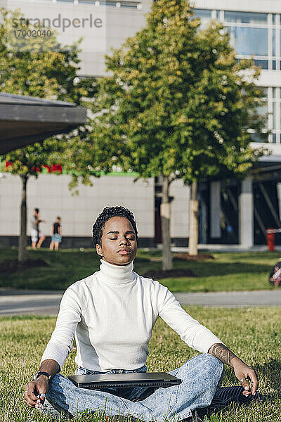 Junge Geschäftsfrau mit Laptop beim Meditieren im Park an einem sonnigen Tag