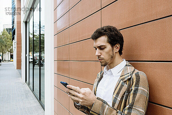 Stylischer junger Mann mit Smartphone vor einem Gebäude stehend