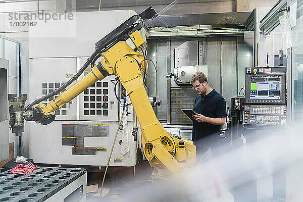Männlicher Fabrikarbeiter verwendet ein digitales Tablet  während er vor Roboterarmen in der Industrie steht