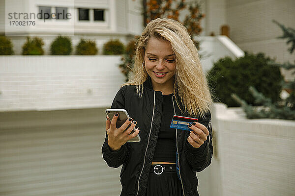Lächelnde blonde Frau  die mit Mobiltelefon und Kreditkarte online einkauft