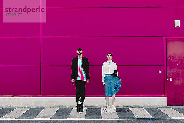 Mann mit Smartphone und Frau mit Kaffee zum Mitnehmen springen vor einer rosa Wand