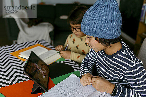 Junge im Gespräch mit einem Lehrer während eines E-Learning-Kurses  während ein männlicher Freund nebenan im Wohnzimmer lernt